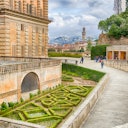 A manicured garden next to a historic building with people walking along a stone pathway; a cityscape and tower are in the background.