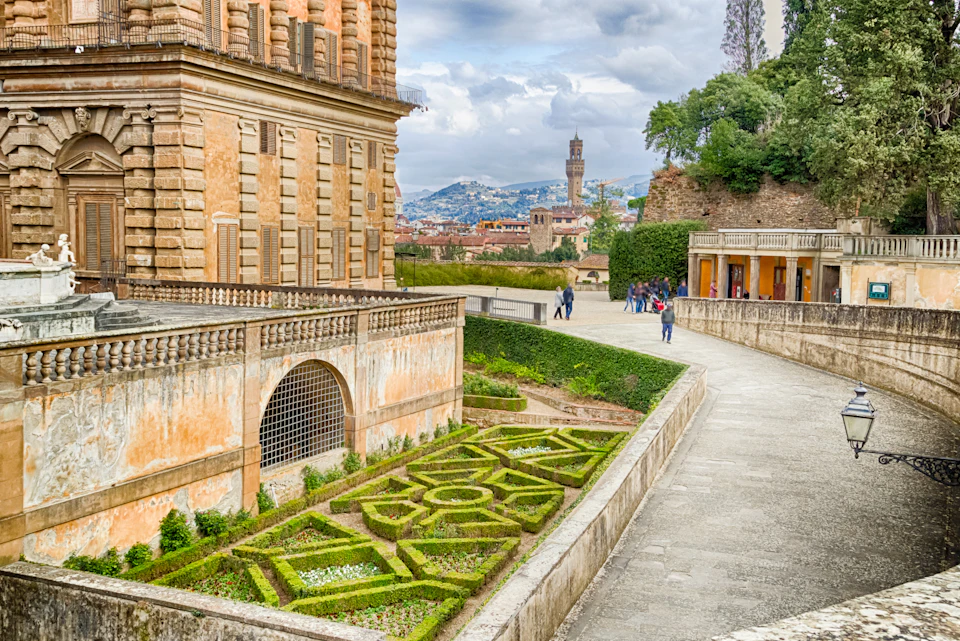 A manicured garden next to a historic building with people walking along a stone pathway; a cityscape and tower are in the background.