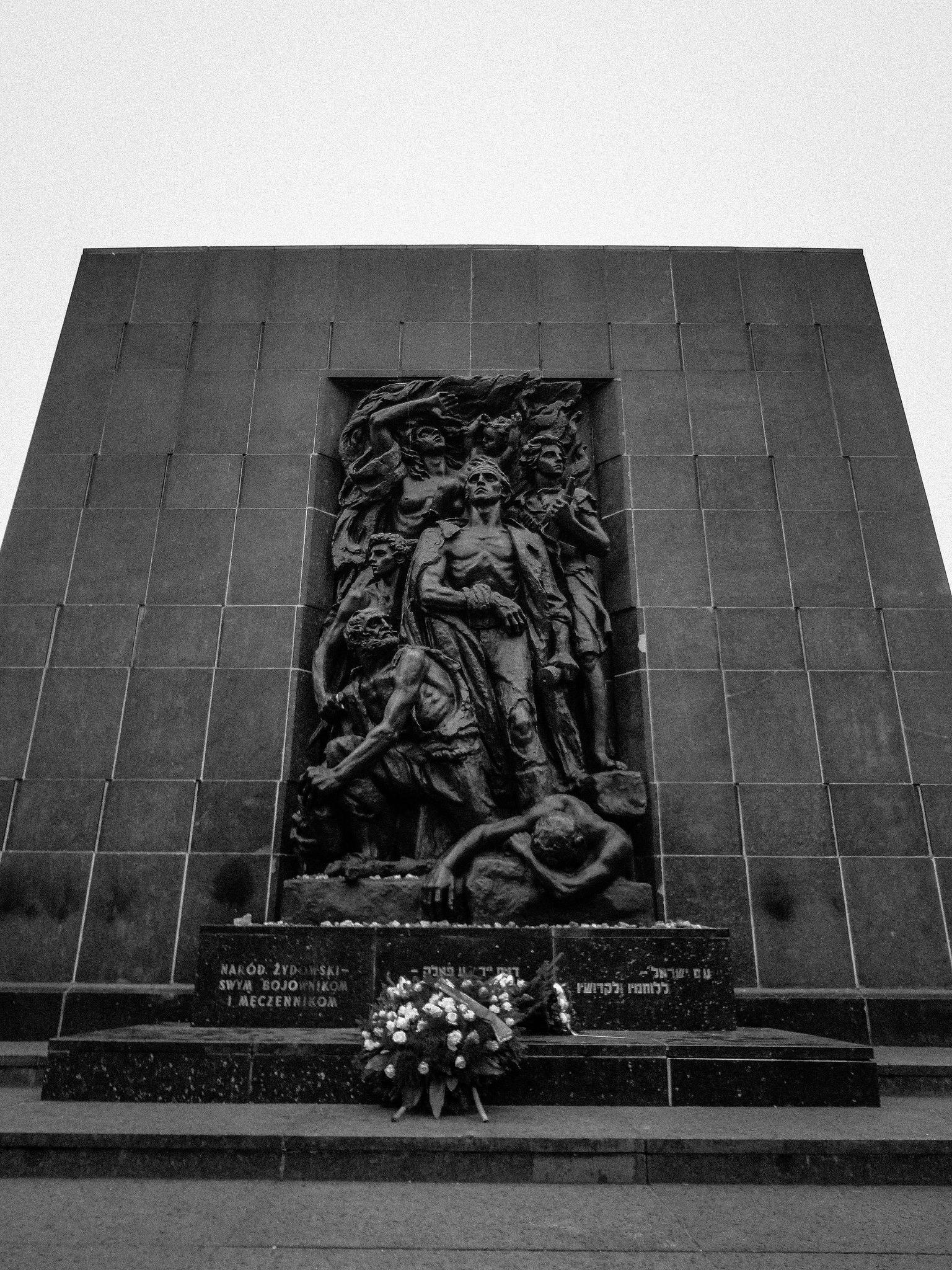 Svartvit skulptur på en monumental vägg med flera mänskliga figurer i en dynamisk pose. Blommig krans vid basen.