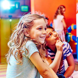 Två unga flickor, den ena leende och den andra tankeväckande, engagerar sig i en interaktiv utställning i en färgglad inomhuslekplats.