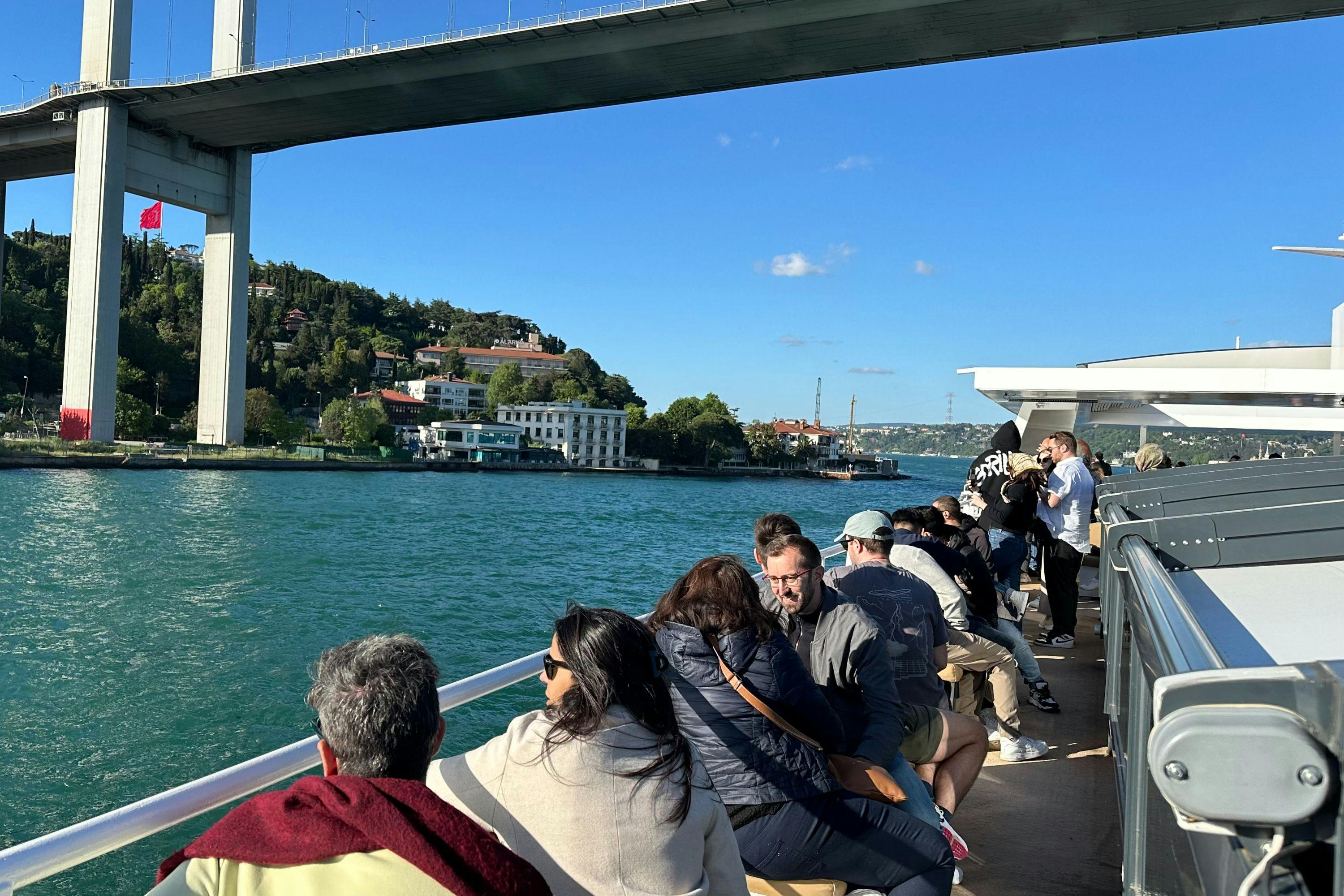 People seated on an open deck boat traveling along a river, with a bridge overhead and greenery on the riverbank.