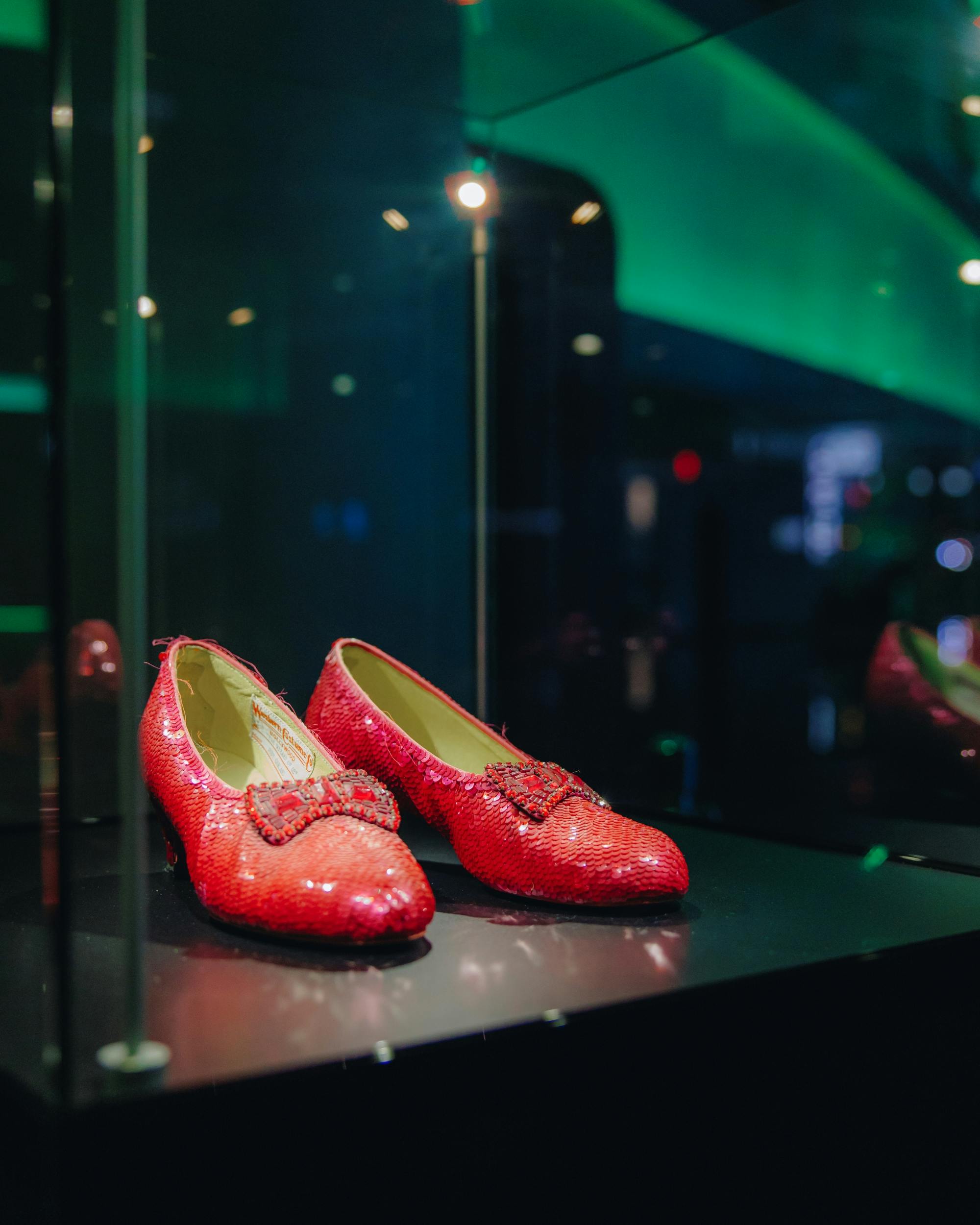 A pair of red, sequined shoes with bows on display in a glass case, under lighting in a museum or exhibit setting.