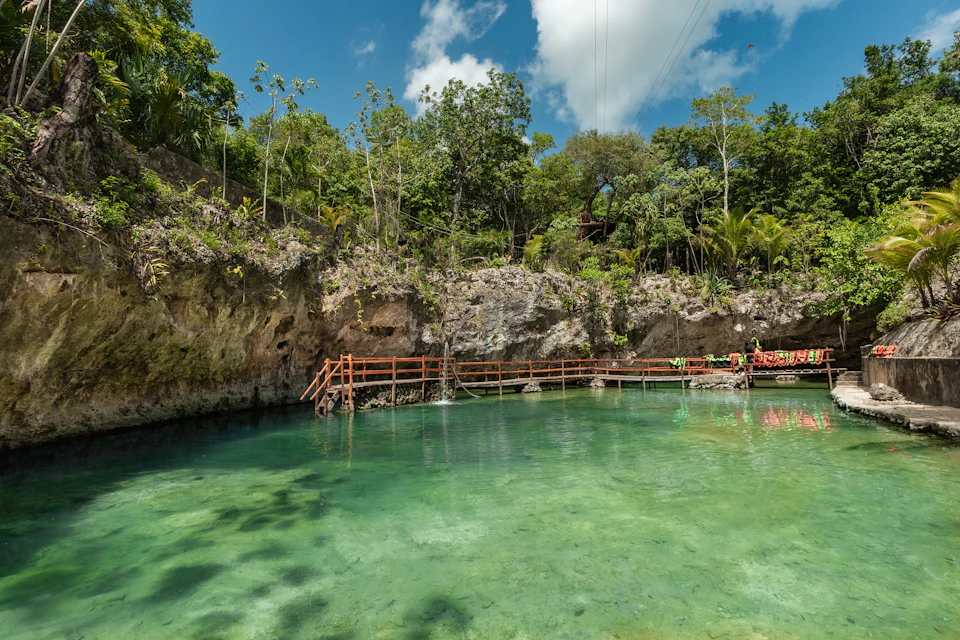 A clear natural pool surrounded by rocky cliffs and dense greenery with a wooden walkway and a few people in the background.