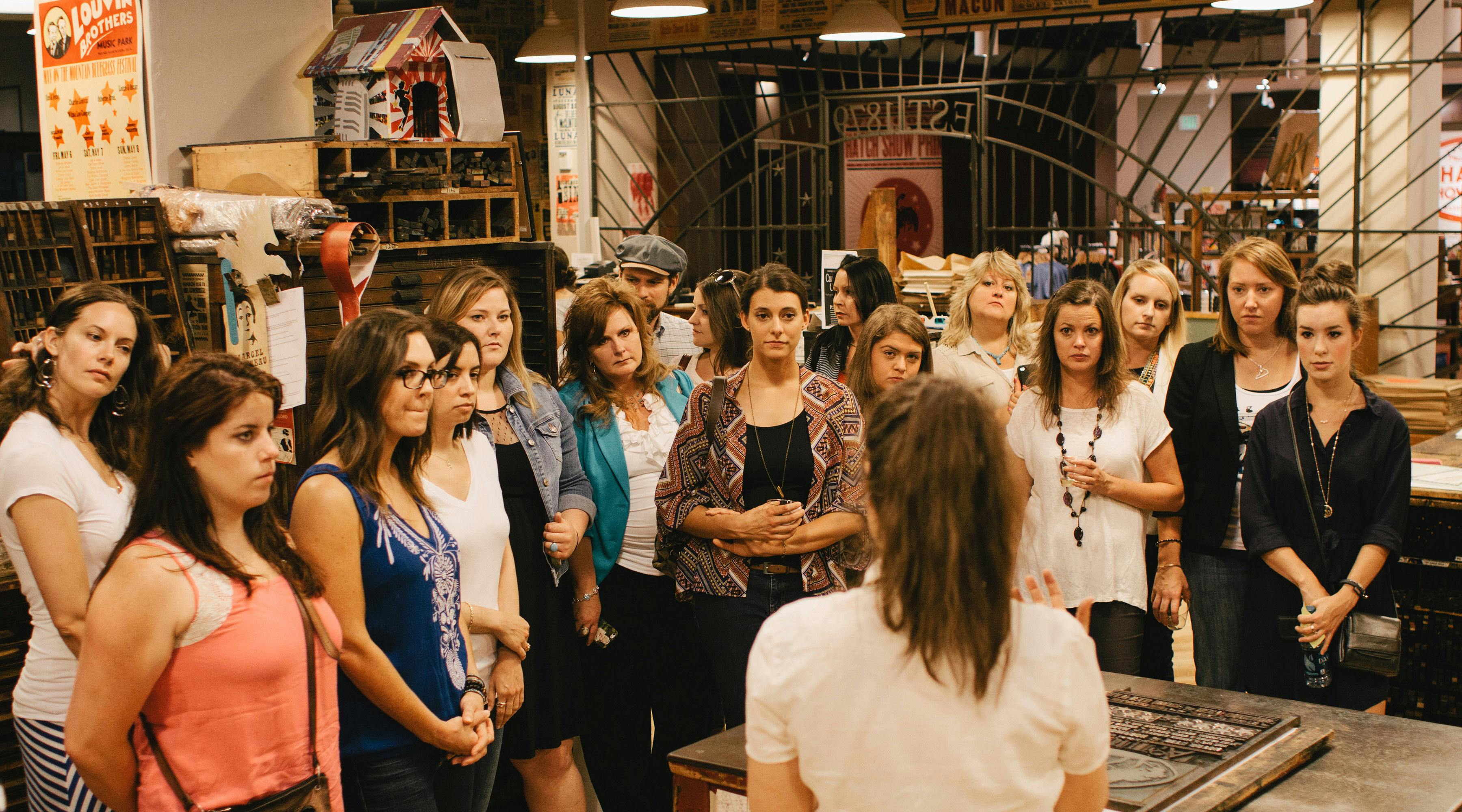 A group of people attentively listening to a speaker in a room with an industrial design and various woodwork pieces visible.