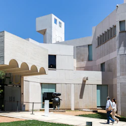 Två personer går förbi en modern byggnad med geometrisk arkitektur, stora fönster och en skulptur nära ingången.