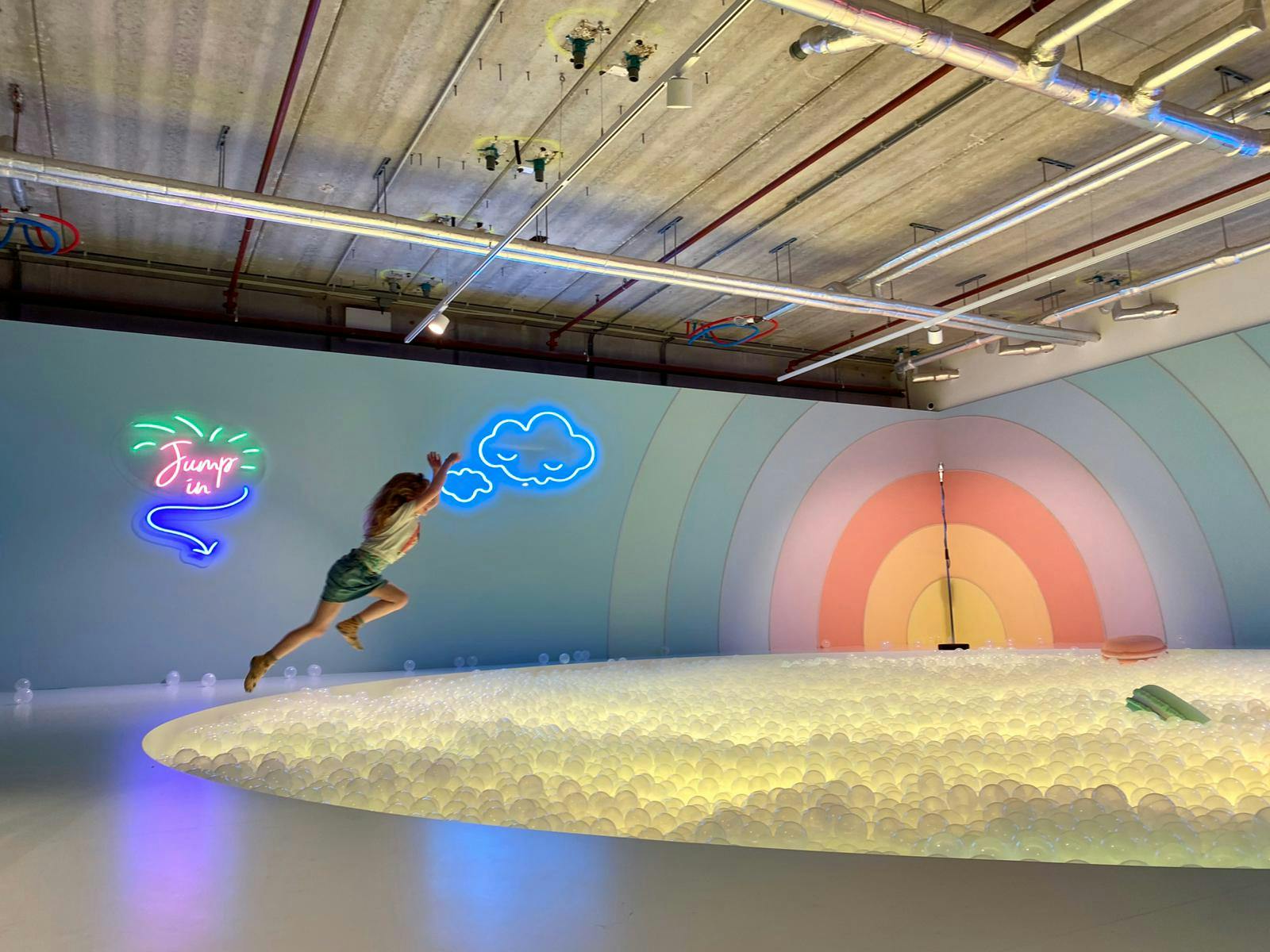 Persona que salta a una fossa de boles il·luminada amb rètols de neó, un mural de l&#39;arc de Sant Martí i canonades de sostre industrials al fons.