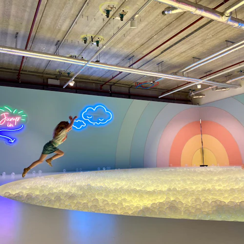 Person som hoppar in i en upplyst bollgrop med neonskyltar, regnbågsväggmålning och industriella takrör i bakgrunden.