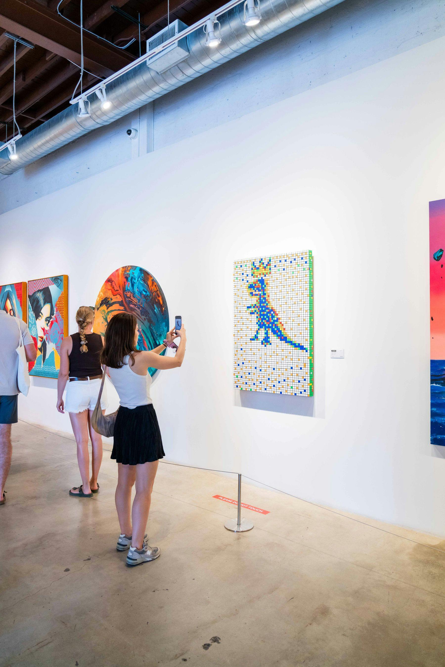 People viewing colorful artworks on a gallery wall; one woman takes a photo with her phone.