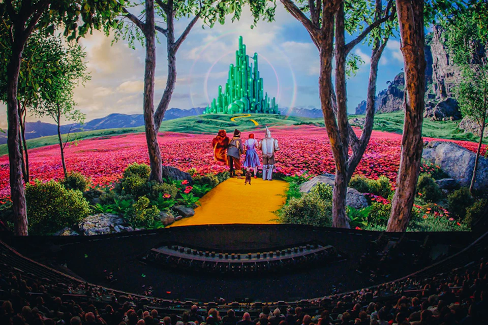 A stage scene depicting characters on a yellow brick road heading towards a green city, surrounded by a colorful landscape.