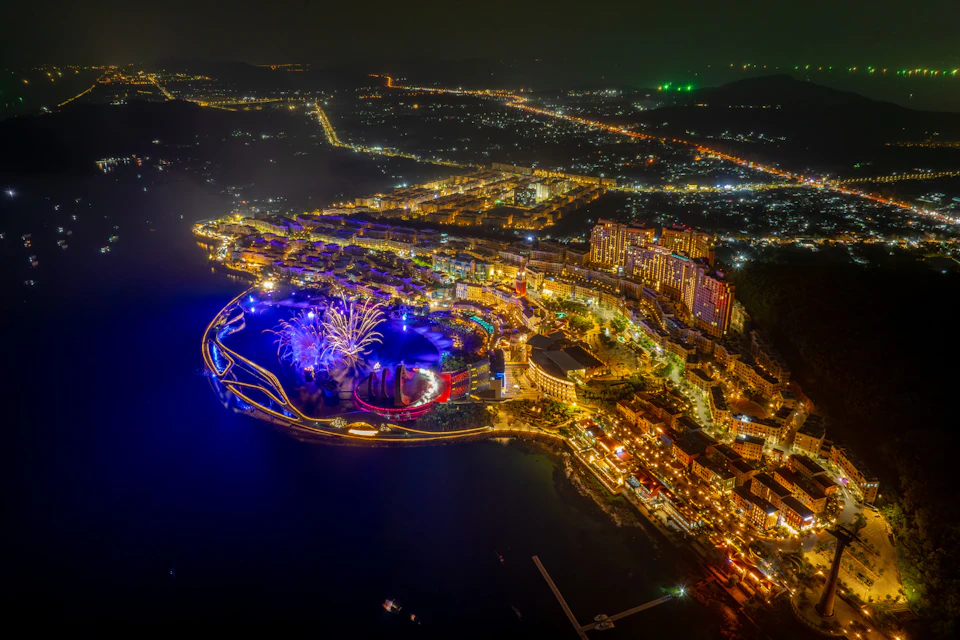 Aerial view of a brightly lit coastal cityscape at night, featuring illuminated buildings and a waterfront area.