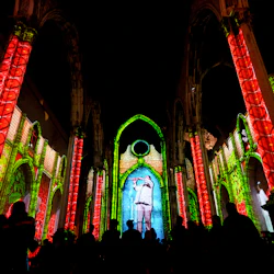 Människor tittar på en ljusprojektion på en katedralens innerväggar, med livfulla röda och gröna mönster och en central figur.
