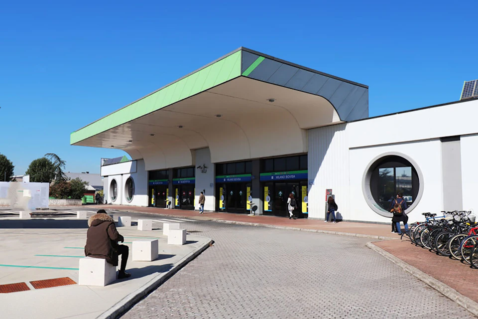 A modern building with a green-roofed entrance, circular windows, and people walking in front under a clear blue sky.