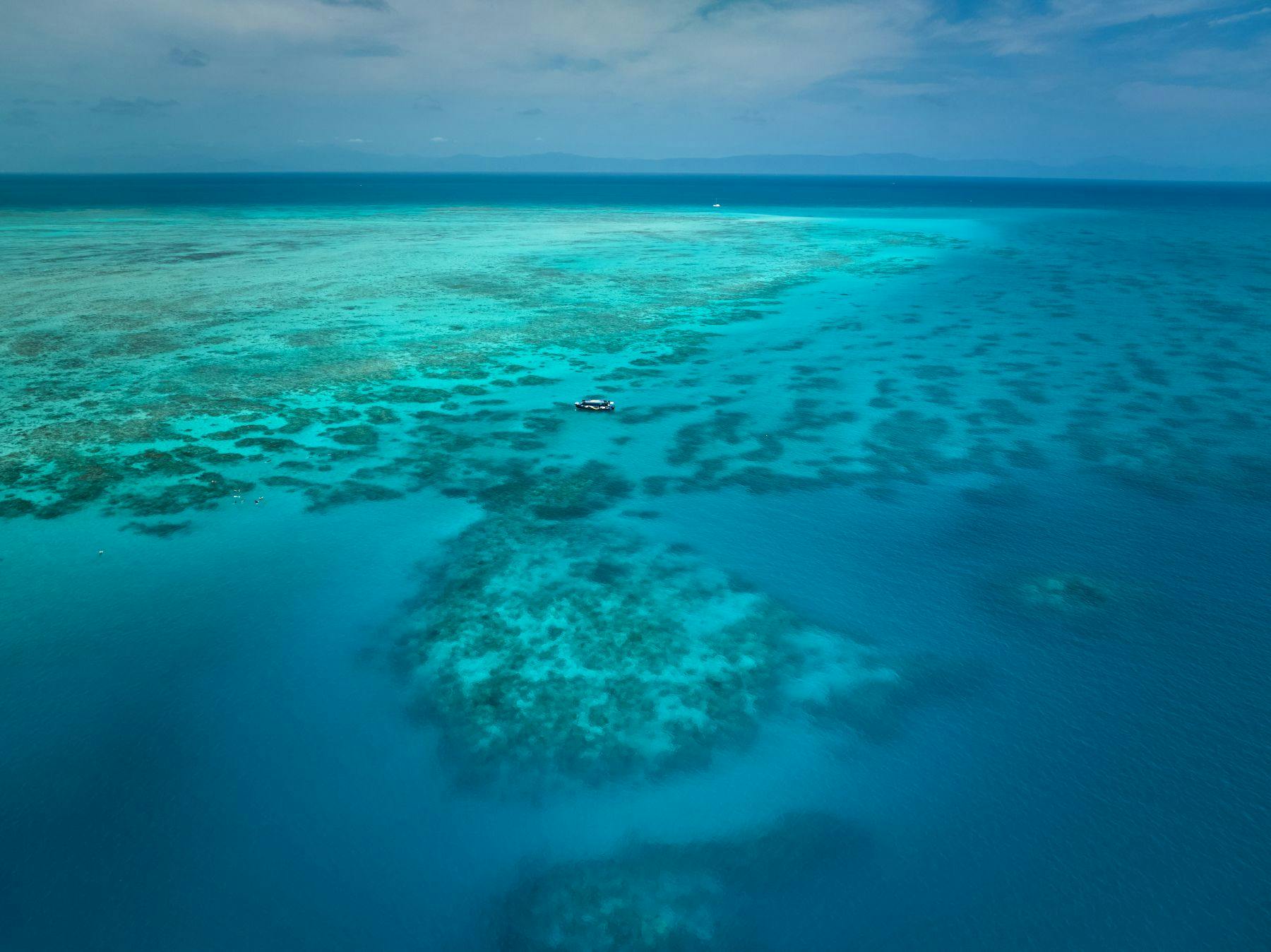 部分的に曇った空の下で遠くの地平線を望む、サンゴ礁の上の澄んだターコイズ色の水の上でのボートの航空写真。