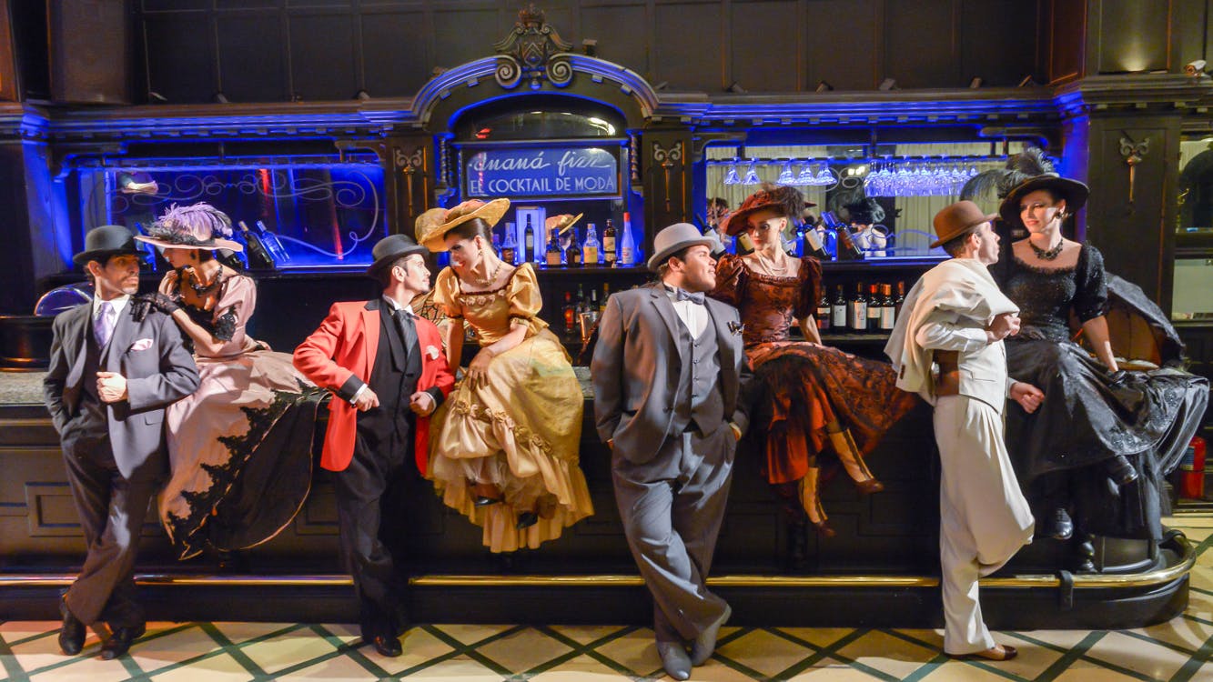 People in vintage attire at a bar; three seated women in hats and dresses chatting with two men in suits and hats.