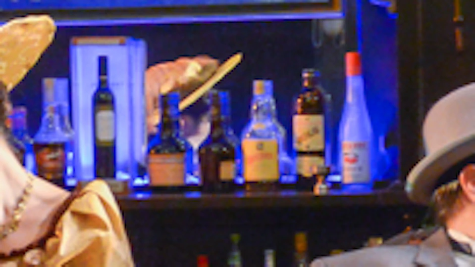 People in vintage attire at a bar; three seated women in hats and dresses chatting with two men in suits and hats.