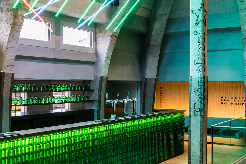 A modern bar interior featuring green neon lighting, a counter made of green bottles, and beer taps in the centre.