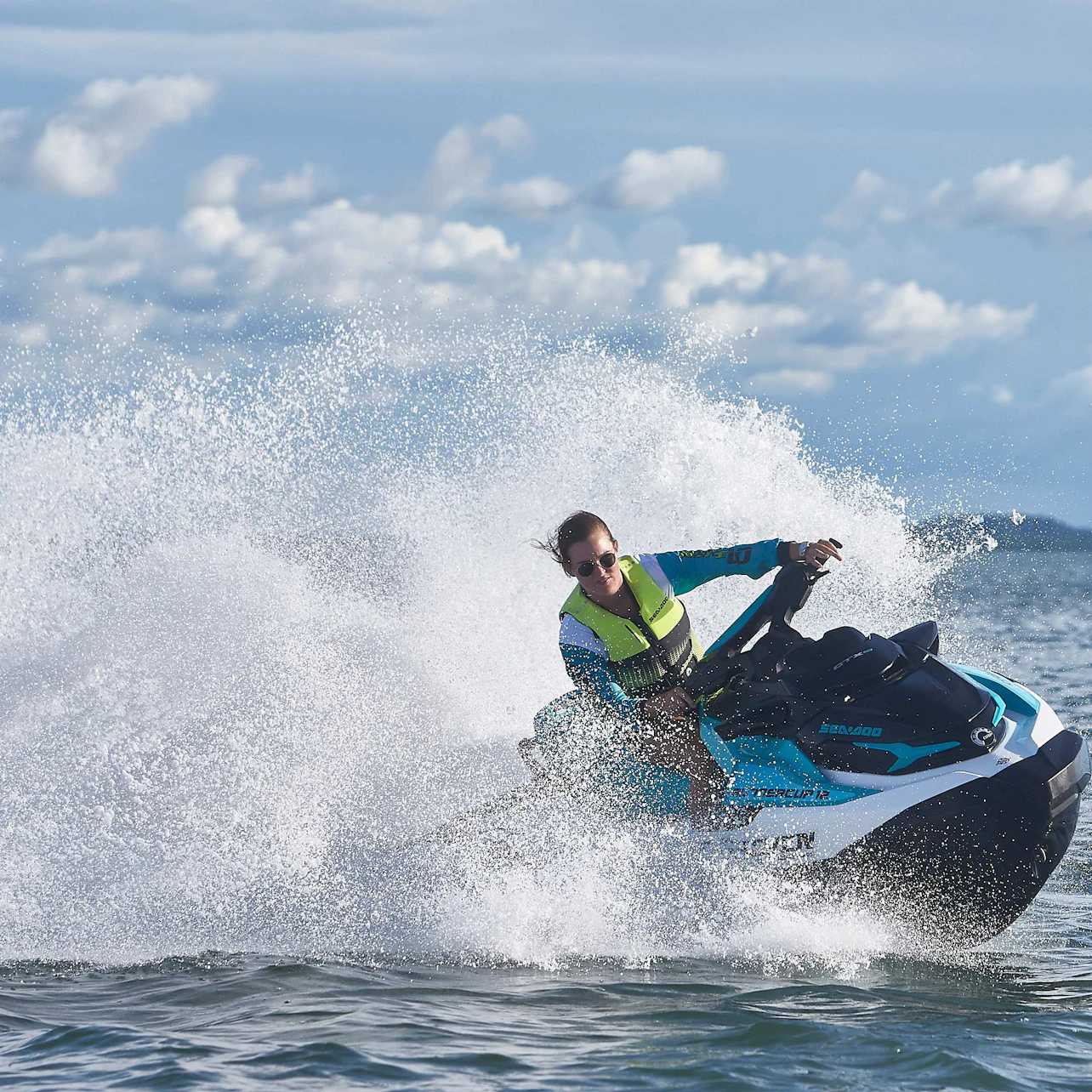 Person riding a blue and black jet ski, creating a large splash on the water under a partly cloudy sky.