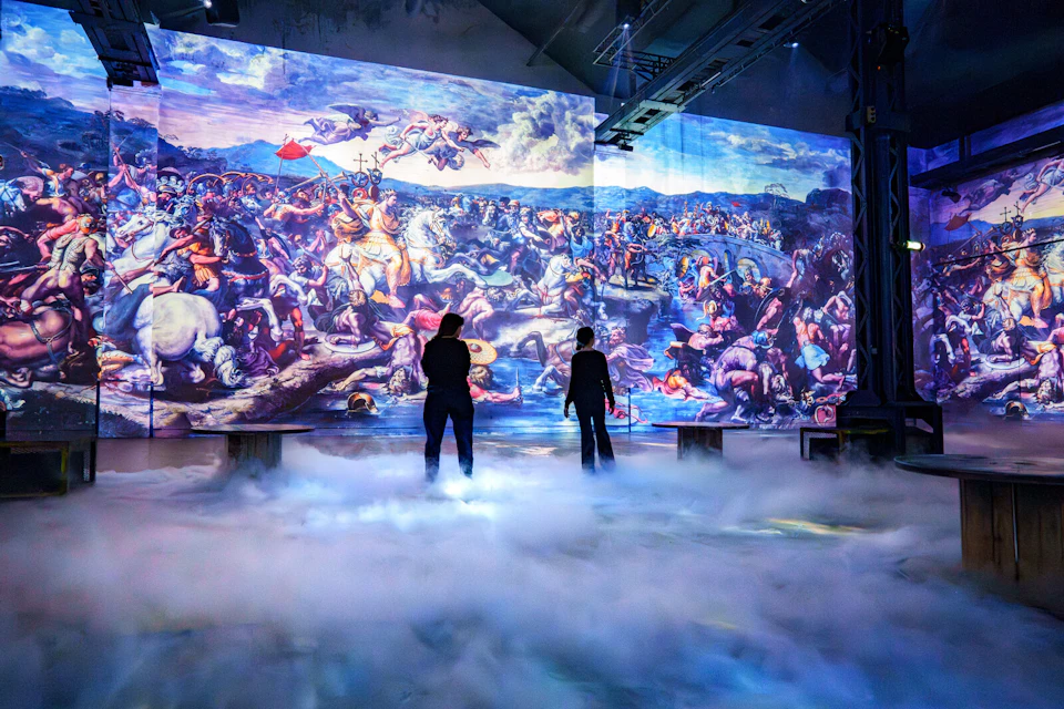 Two individuals stand in a misty room observing a large digital projection of a Renaissance-style battle painting.