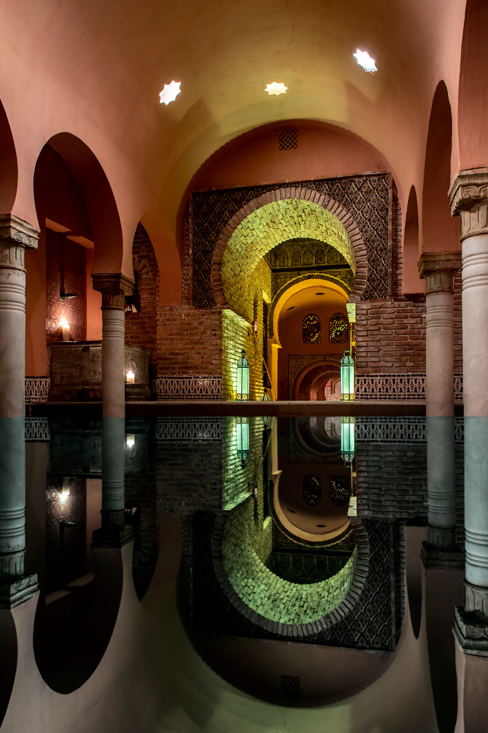 Interior of a dimly lit bathhouse with arches, columns, brick walls, and a reflective water surface.