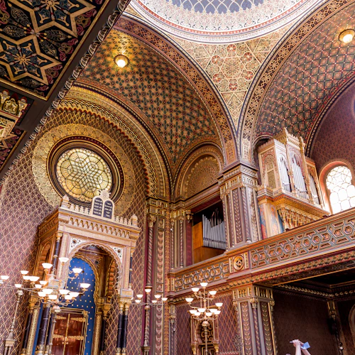 Invecklad interiör med utsmyckade tak, välvda fönster, ljuskronor och ett stort glasmålningsfönster, med rika mönster och färger.