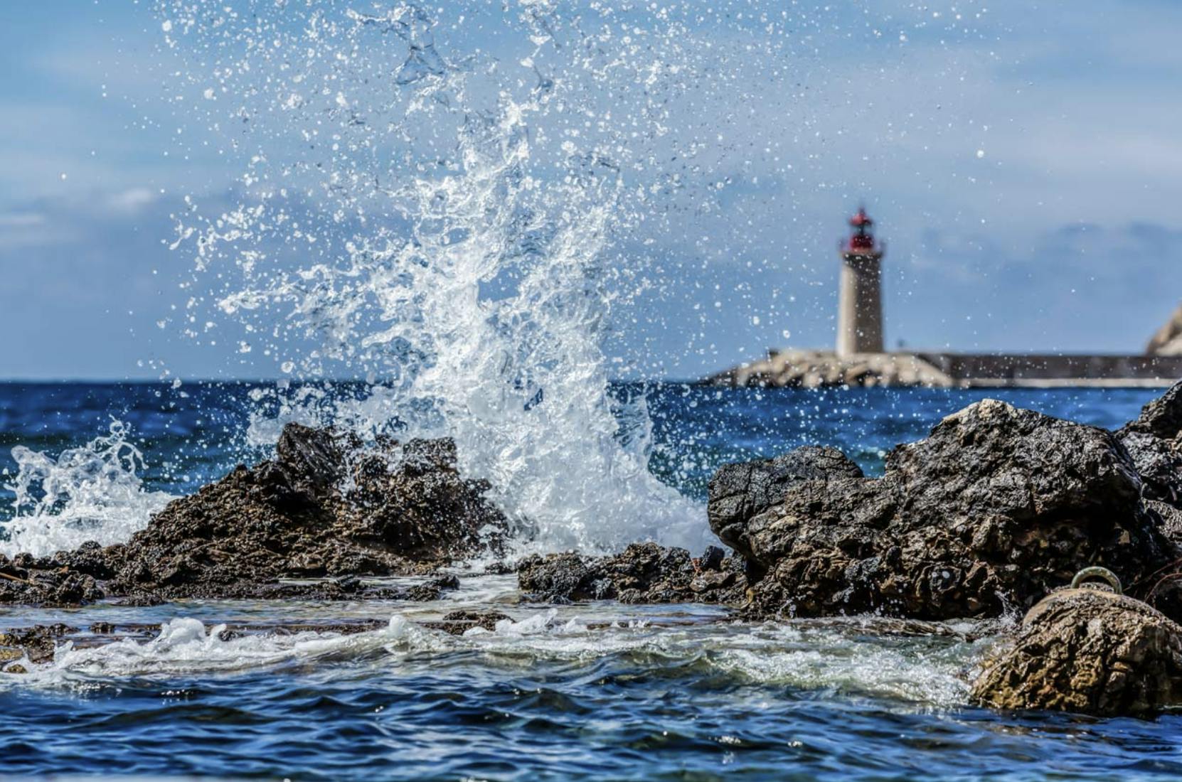 Wellen schlagen gegen eine felsige Küste mit einem entfernten Leuchtturm auf einer kleinen Insel unter einem teilweise bewölkten Himmel.