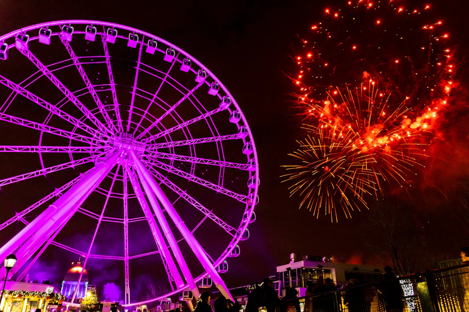 A lit ferris wheel glows pink against the night sky, while bright fireworks explode in the background. People are gathered below.