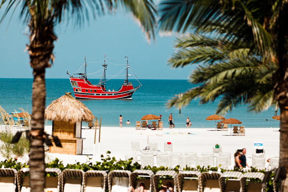 People on a sunny beach with lounge chairs and umbrellas. A red pirate ship is floating in the calm, blue ocean in the background.