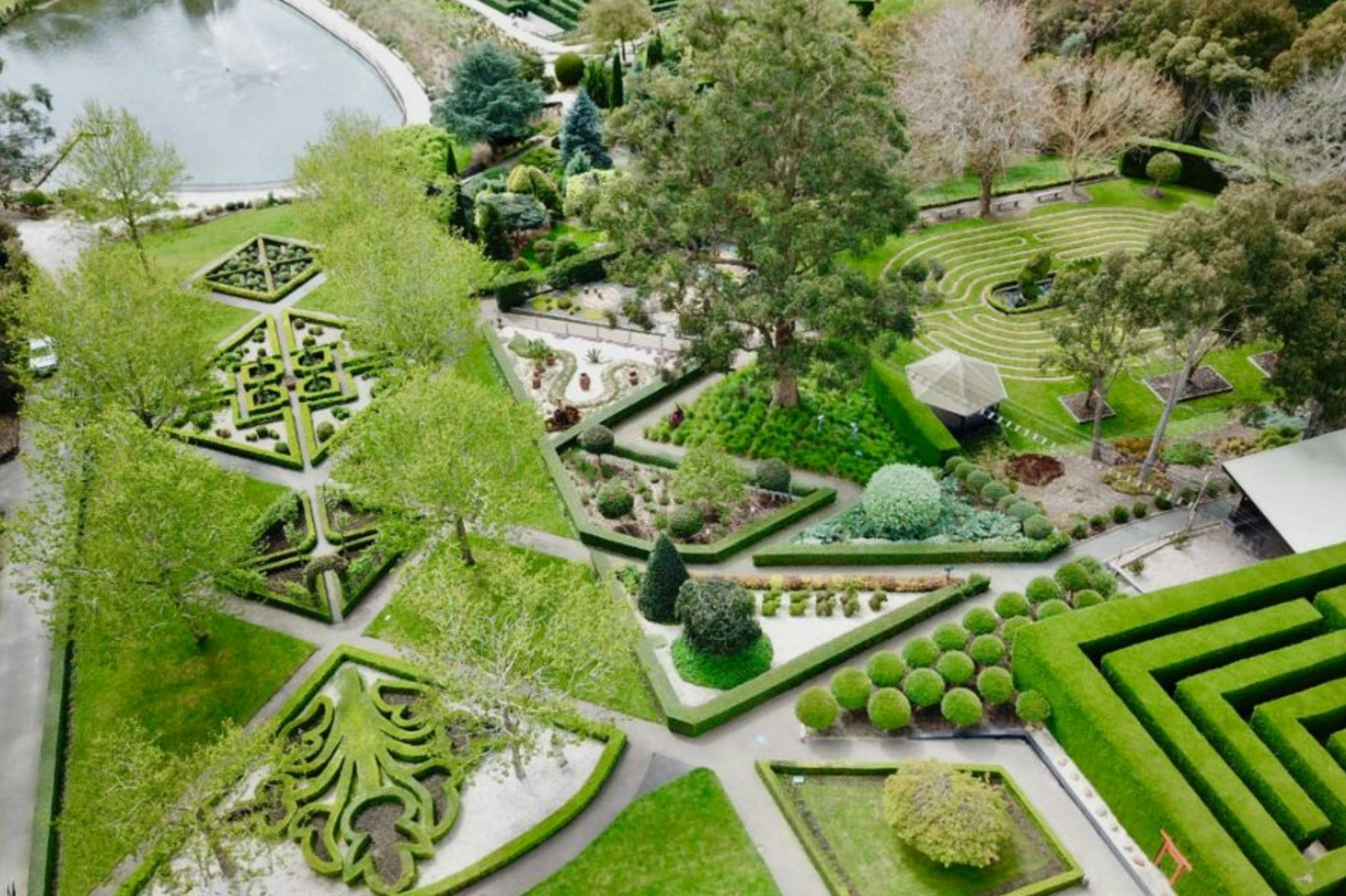 Vue aérienne d'un jardin à la française avec des haies géométriques, des allées et un étang entouré d'arbres.