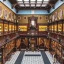 An opulent, historical gallery with intricate architecture, two levels of framed portraits, and display cabinets surrounding a tiled floor.