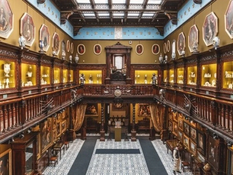 An opulent, historical gallery with intricate architecture, two levels of framed portraits, and display cabinets surrounding a tiled floor.