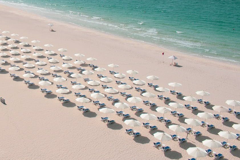 Rows of white beach umbrellas and lounge chairs on a sandy beach beside turquoise ocean waves.