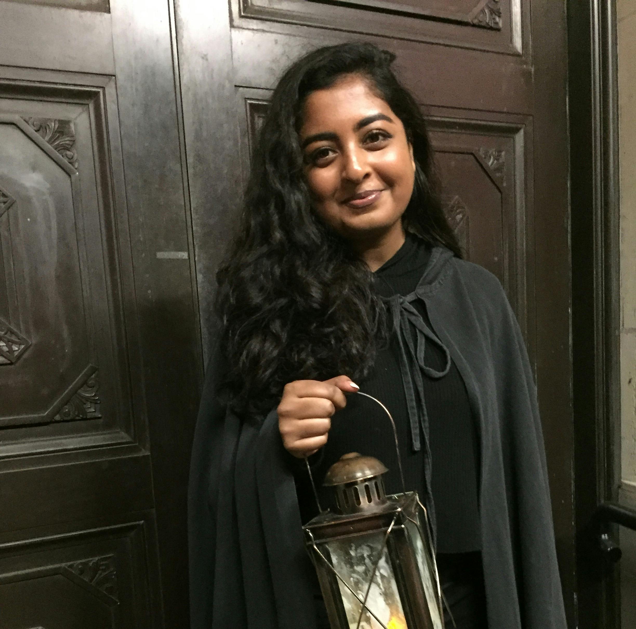 一位留着深色波浪头发的人拿着灯笼，站在一扇带有装饰板的大木门前。