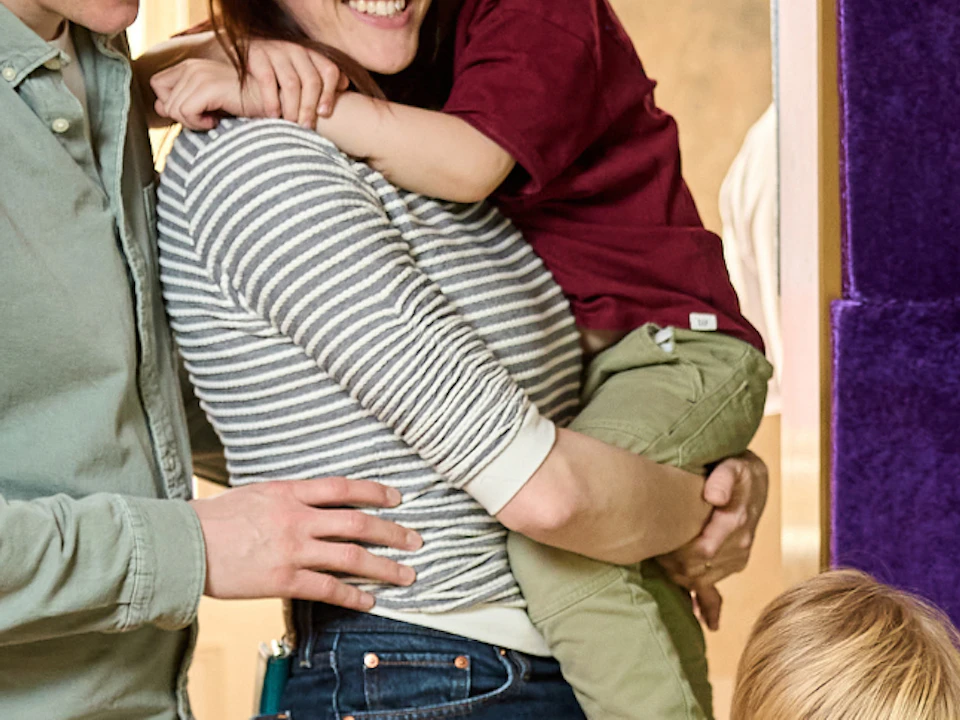 A family of four gathers around a small piano. The parents smile while a boy plays the piano and another child is held by the mother.