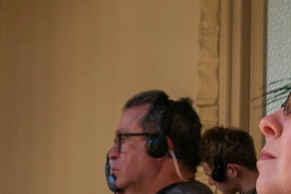 A tour guide gestures to a window while speaking to a group of people in a room with light streaming in.
