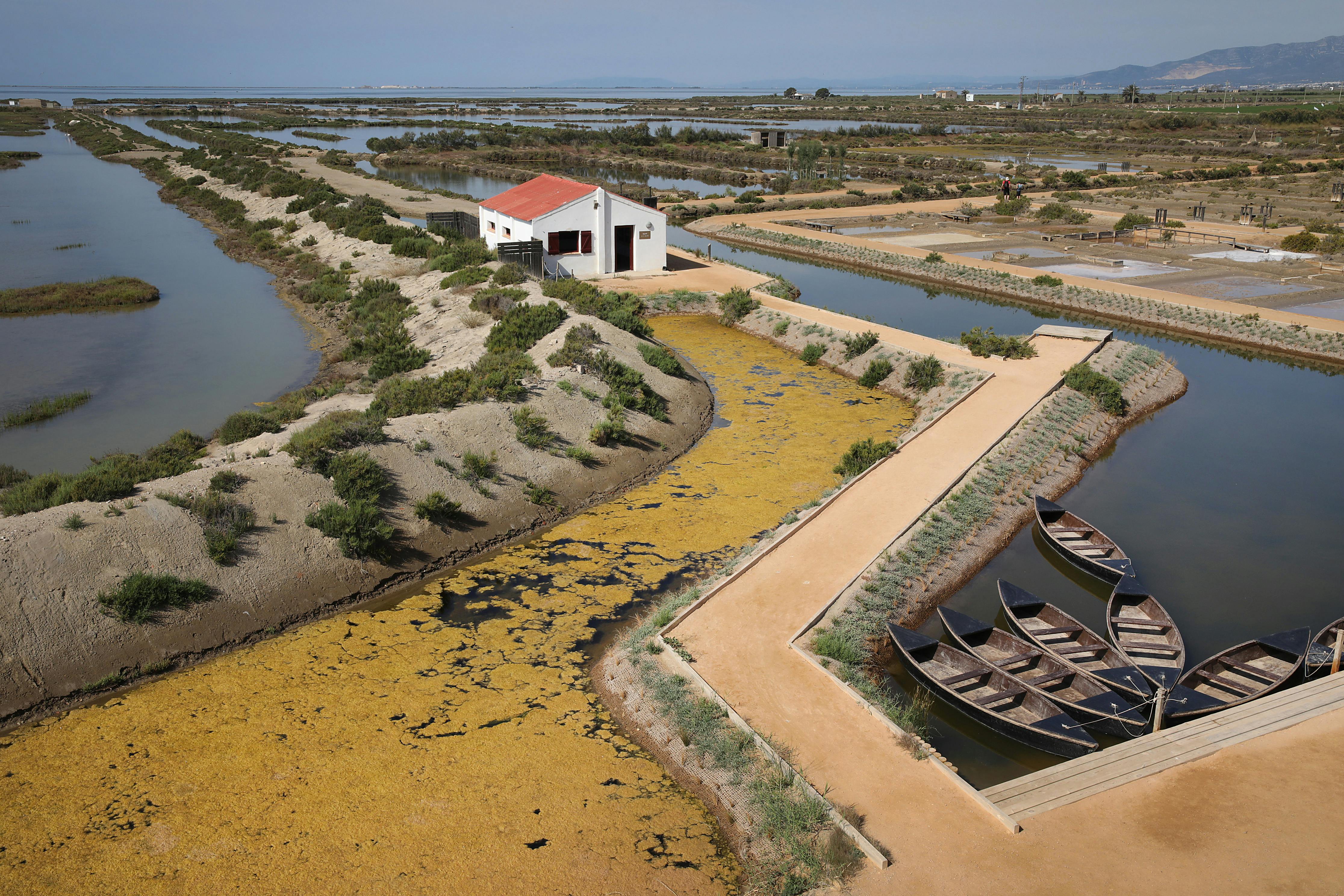 Ένα λευκό σπίτι με κόκκινη στέγη βρίσκεται ανάμεσα σε κανάλια νερού με μικρές βάρκες αγκυροβολημένες δίπλα σε ένα μονοπάτι σε ένα ελώδες τοπίο.