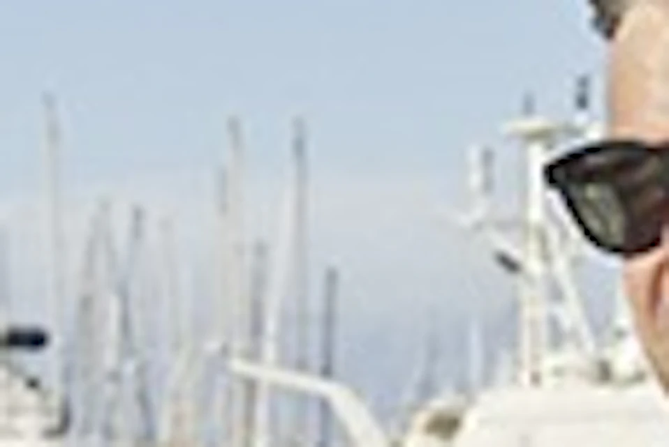 Four men riding bicycles on a dock, with sailboats and a clear blue sky in the background.