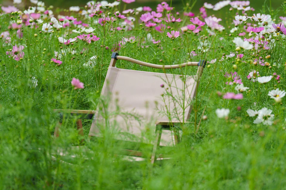 A beige foldable chair surrounded by tall grass and blooming pink and white flowers in a garden setting.