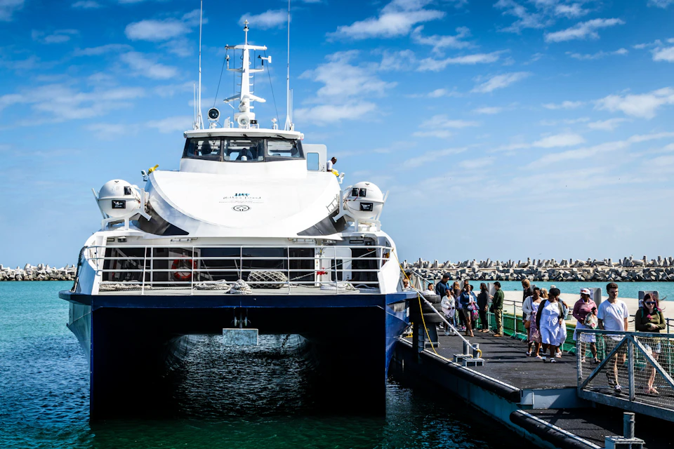People boarding a large white ferry docked at a pier under a partly cloudy blue sky.