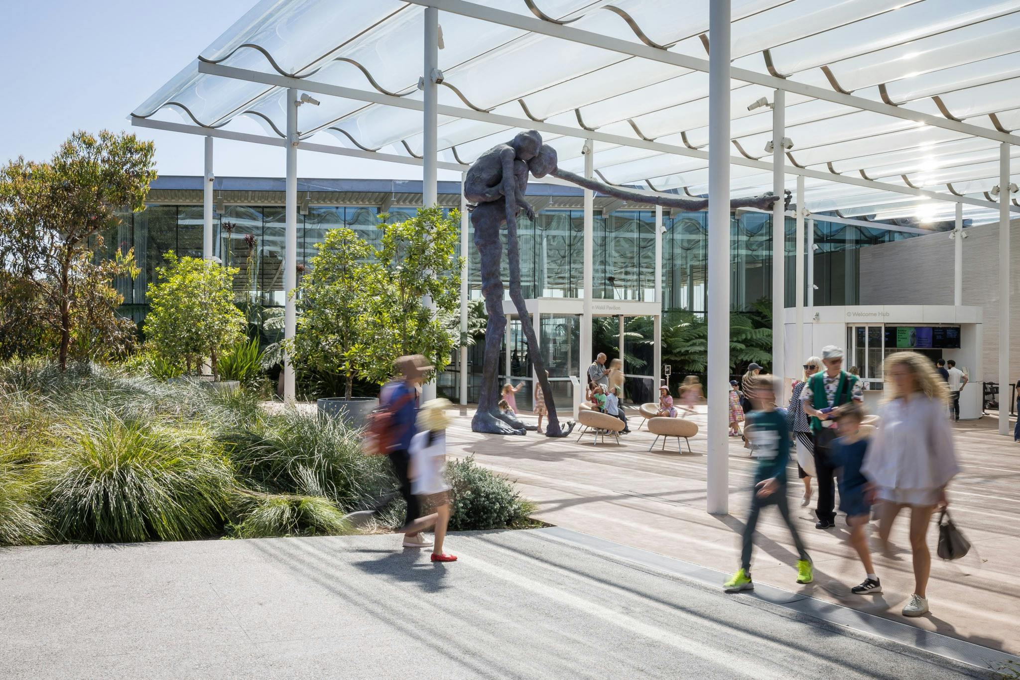 A modern outdoor plaza with a large sculpture of a figure leaning on a stick, surrounded by people and greenery.