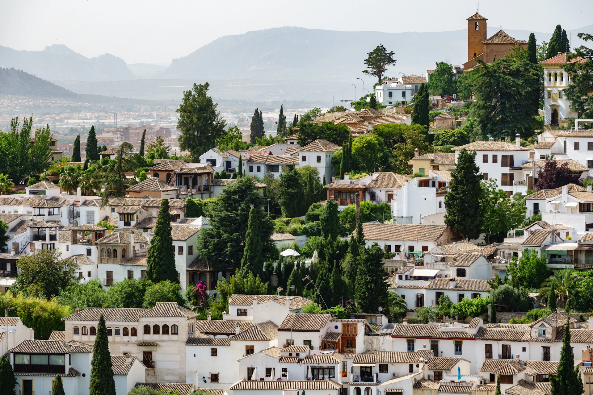A hillside village with white buildings, terracotta roofs, and trees, with mountains visible in the background.