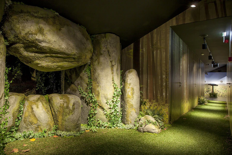 A room with artificial grass, large rocks with vines, and a wallpaper of a forest scene. A door is visible on the right.