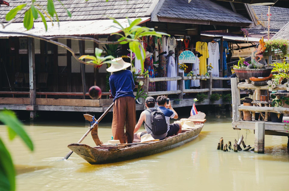 A small wooden boat with three people is floating on a canal near a wooden market building displaying colorful clothing.