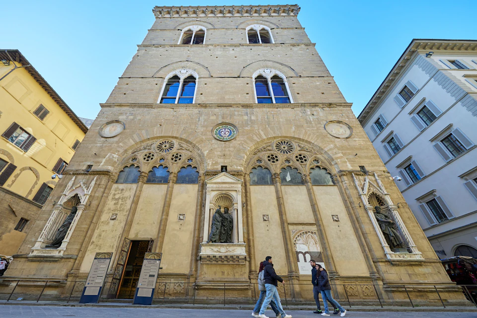 A historic building with ornate architecture and statues, with three people walking in front under a clear blue sky.