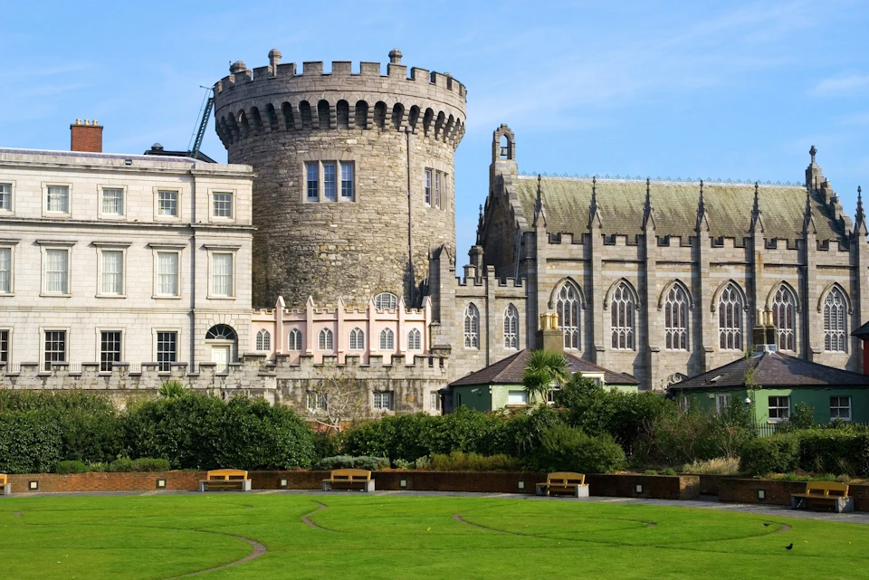 Historic stone castle and church with arched windows, surrounded by greenery and benches, under a clear blue sky.