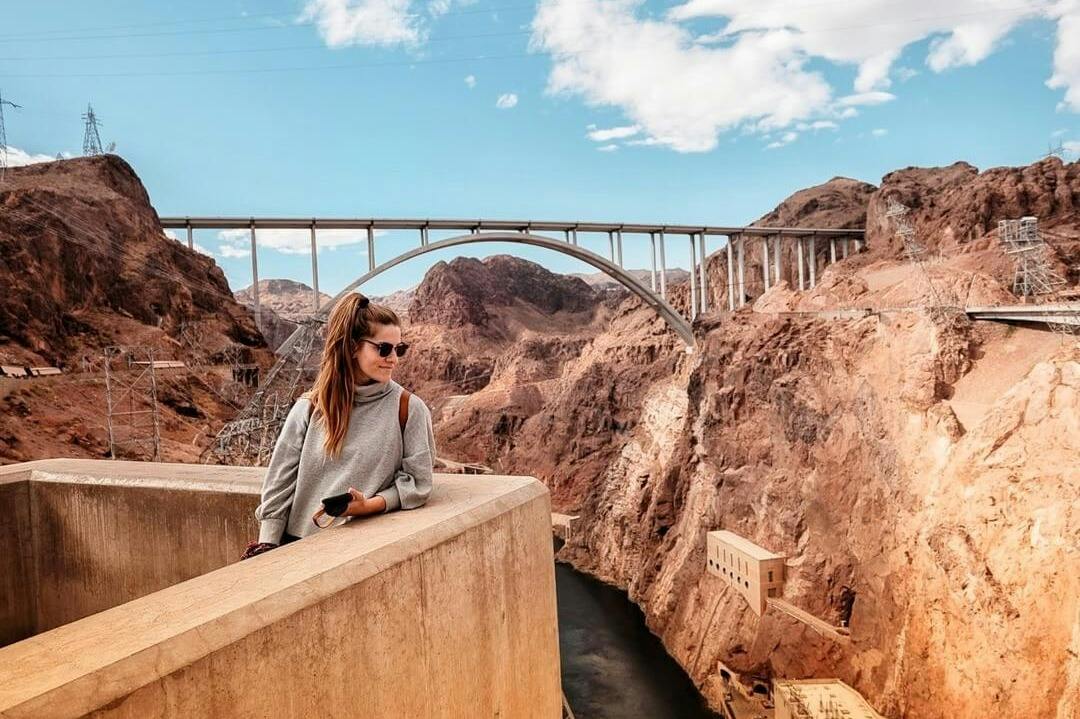 A woman in a gray sweater stands on a concrete balcony, with a bridge and rocky canyon landscape in the background.