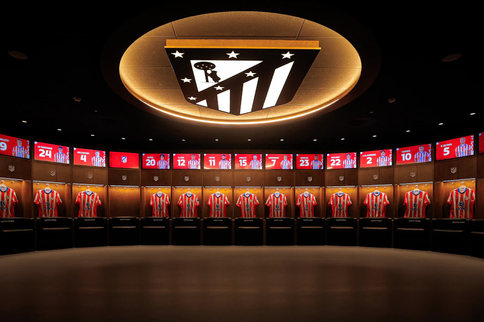A locker room featuring red and white striped soccer jerseys displayed under bright lighting, with a large logo on the ceiling.