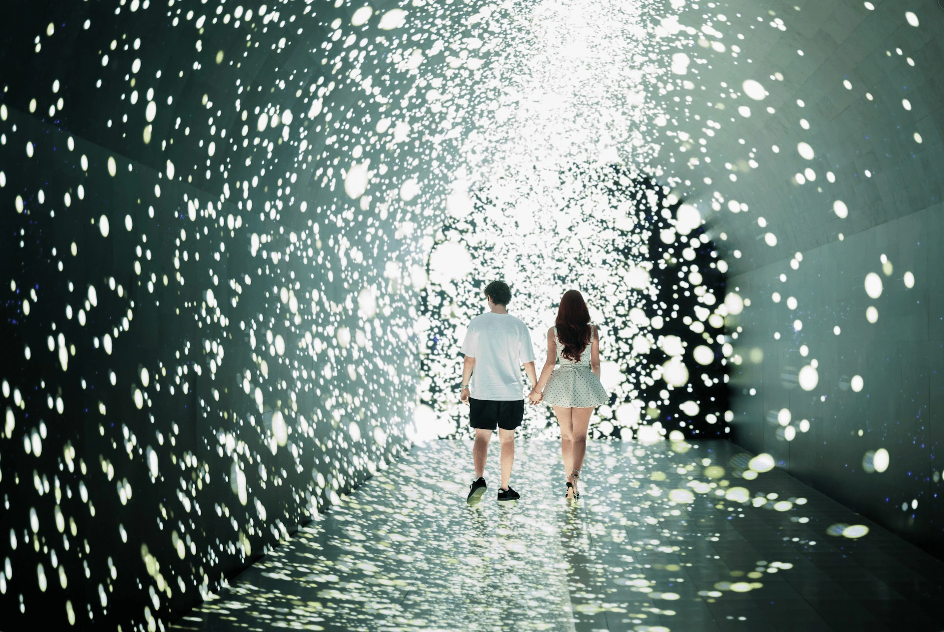 A couple walking hand-in-hand through a tunnel filled with bright, glowing spots resembling stars or lights.