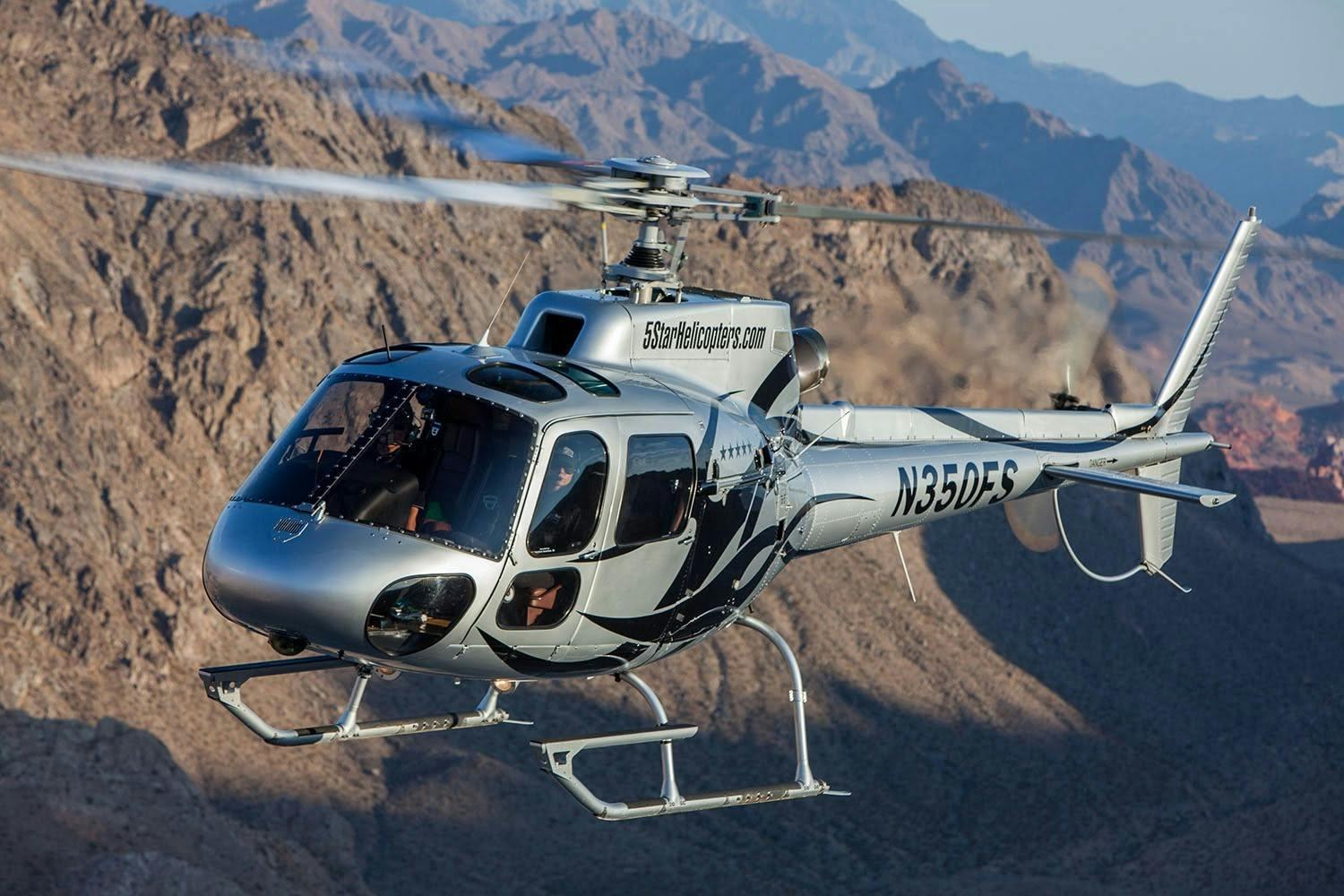 A silver helicopter in flight with mountains in the background.