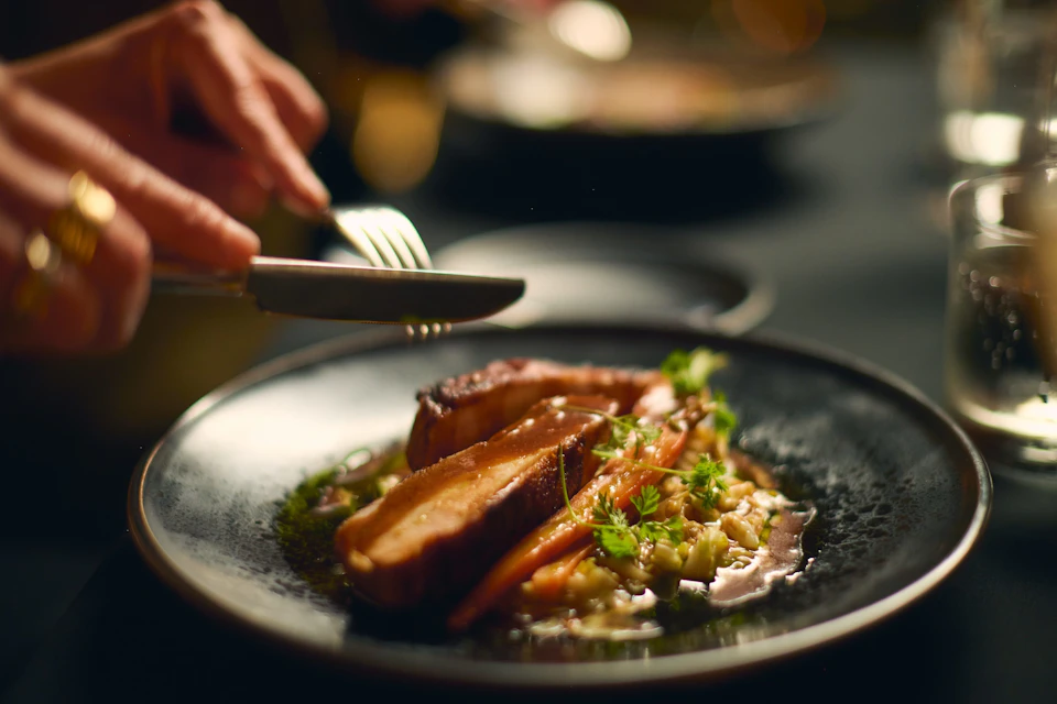 A plated dish of cooked meat, vegetables, and garnish, with a person using a knife and fork in the background.