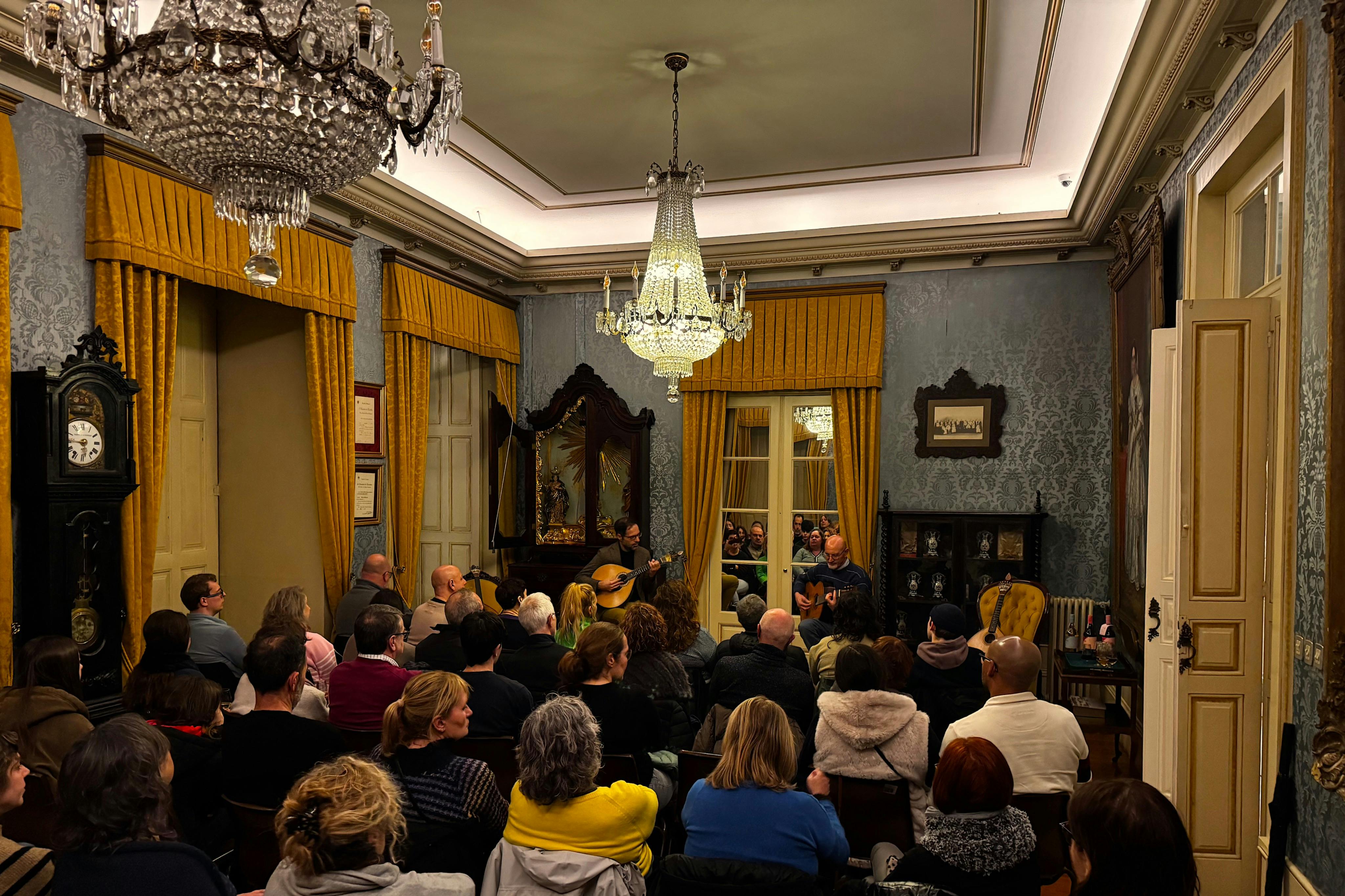 A group of people sitting in a room watching a performance under a chandelier, with ornate decor and yellow curtains.